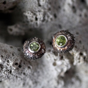 Op de afbeelding: Een paar zilveren oorbellen met groene edelstenen, gezet in een getextureerd, organisch ontwerp. De oorbellen worden getoond op een wit, getextureerd oppervlak.