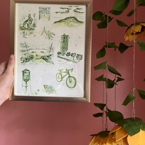 以下が含まれることがあります： 自転車、給水塔、建物、丘、ウサギのある町の水彩画の額縁。画像には「Coinsett Hotel」というテキストが表示されています。