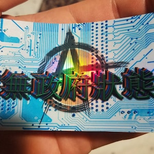 May include: A sticker with a blue and white circuit board design. The sticker features a holographic, rainbow-colored anarchy symbol with the text "無政府 崩壊 覚醒" in black, green, and orange.