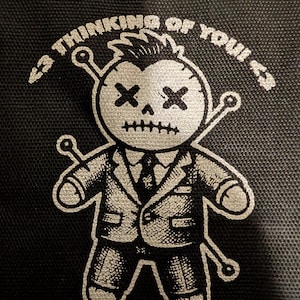May include: A black graphic featuring a voodoo doll in a suit with crossed-out eyes and mouth. The text "I'M THINKING OF YOU" arches above the doll in a white, stylized font. The doll has pins through its head and limbs.