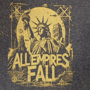 May include: A gold graphic print on a dark grey t-shirt featuring the Statue of Liberty and the text "All Empires Fall".