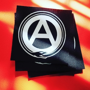 May include: Black and white patch with an anarchy symbol inside a circle. The symbol is surrounded by two hands.