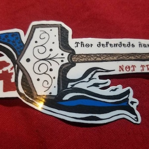 May include: A white sticker with a black and white illustration of Thor's hammer, Mjolnir. The hammer is depicted with a blue and black cloud behind it. The text "Thor defends humanity NOT TROLLS!" is written in black on the sticker.