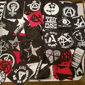 May include: A collection of black and white patches with various symbols and slogans, including anarchy, peace, and anti-establishment messages. The patches are made of fabric and have a distressed look. Some of the patches are red and white.