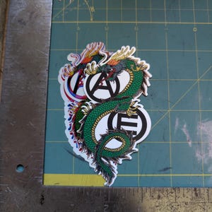May include: A collection of stickers featuring a green dragon design with red, yellow, and blue accents. The stickers also include the letters "A" and "E" in a circular design. The stickers are on a green cutting mat.