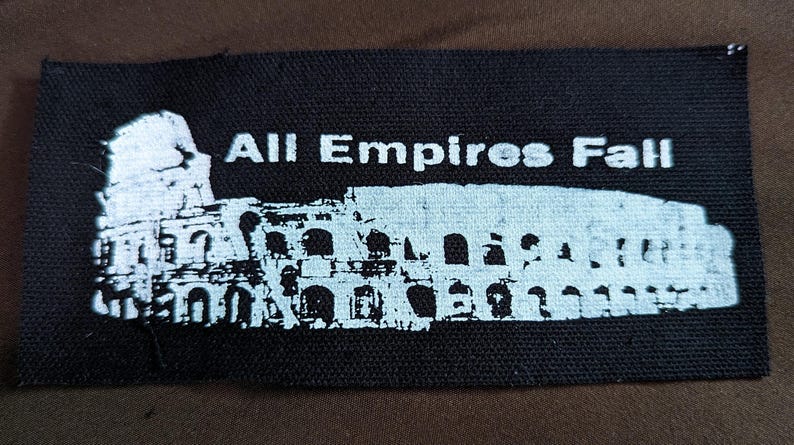 Puede incluir: Parche negro con texto blanco que dice "All Empires Fall" y una silueta blanca del Coliseo.