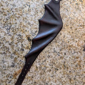 May include: A dark brown leather wrist cuff with a bat wing design. The cuff features a buckle closure and a strap with multiple holes for adjustment. The leather has a textured appearance.
