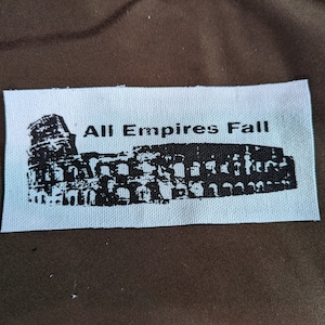 Puede incluir: Parche de tela blanco con tinta negra que representa el Coliseo y el texto "All Empires Fall".