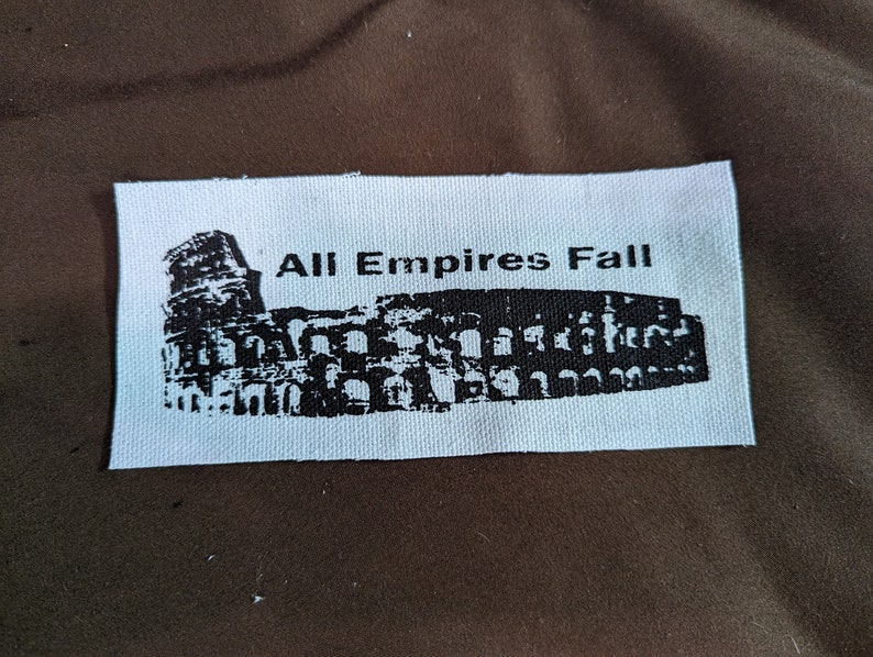 Puede incluir: Parche de tela blanco con tinta negra que representa el Coliseo y el texto "All Empires Fall".