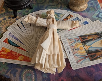 May include: A light beige corn husk doll with a tied waist and outstretched arms resting on a fan of tarot cards. To the left is a small black three-legged cast iron cauldron with a lid, and to the right is a speckled tan stone mortar and pestle.