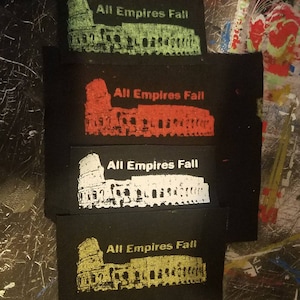 Puede incluir: Cuatro parches de tela negra con el texto "All Empires Fall" y una silueta blanca del Coliseo en diferentes colores: verde, rojo, blanco y dorado.
