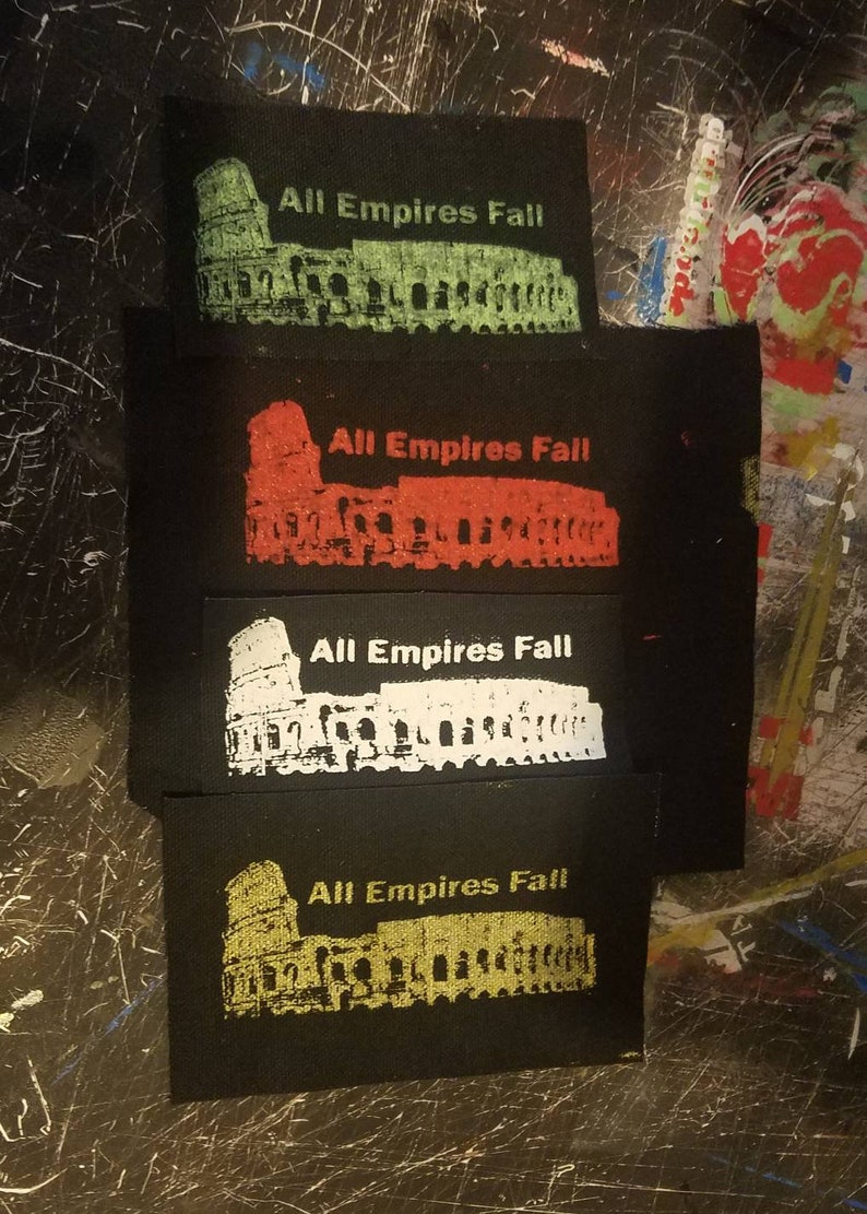 Puede incluir: Cuatro parches de tela negra con el texto "All Empires Fall" y una silueta blanca del Coliseo en diferentes colores: verde, rojo, blanco y dorado.