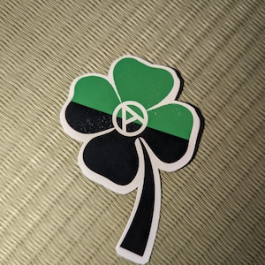 May include: A black and green four-leaf clover sticker with an anarchy symbol in the center. The sticker has a white outline.
