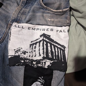 May include: A pair of blue denim jeans with a black and white patch sewn on the front. The patch features a black and white image of a building with the text "ALL EMPIRES FALL" above it.