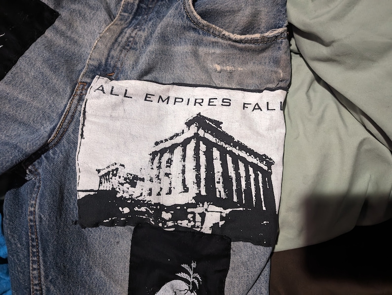May include: A pair of blue denim jeans with a black and white patch sewn on the front. The patch features a black and white image of a building with the text "ALL EMPIRES FALL" above it.