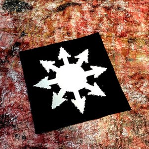 May include: A black square patch featuring a white, stylized eight-pointed starburst design. The starburst has sharp, arrow-like points radiating outwards. The background has a textured, multi-colored appearance with shades of red, brown, and white.