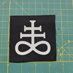 May include: White symbol of a cross with an infinity symbol at the bottom on a black fabric patch.