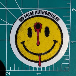 May include: A round sticker featuring a yellow smiley face with a bullet hole and dripping blood. The text "NO FALSE AUTHORITIES!!" is printed around the edge of the smiley face in black.