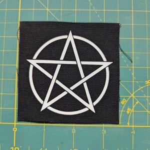 May include: Black square patch featuring a white pentagram design within a circle. The patch is made of a textured fabric. The pentagram is a five-pointed star, a symbol often associated with occult or alternative spiritual practices.