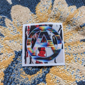 May include: A square patch with a colorful design featuring a stylized "A" inside a circle. The design incorporates a spectrum of colors, including red, blue, and yellow. The patch is set against a textured, patterned background.