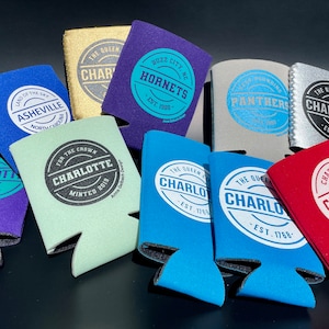 May include: Assortment of 10 colorful can coolers with various logos and text. The logos include "Charlotte Checkers", "Charlotte Hornets", "Asheville", "The Queen City Charlotte", and "For the Crown Charlotte".