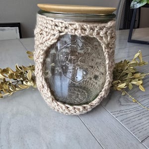 May include: A glass jar with a wooden lid and a crocheted beige cover. The jar is sitting on a wooden table.