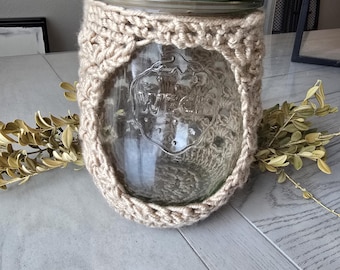 Op de afbeelding: Een glazen pot met een houten deksel en een gehaakte beige hoes. De pot staat op een houten tafel.