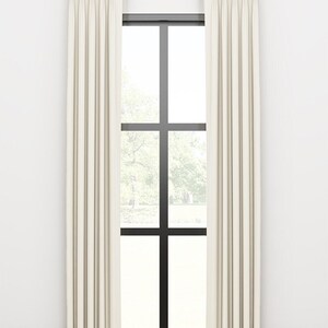 May include: A pair of off-white curtains with pleats hanging from a gold curtain rod in front of a window with black trim.
