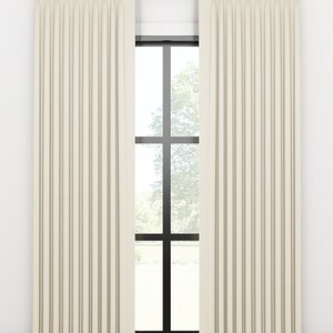 May include: A pair of off-white curtains with a subtle striped pattern hang from a gold rod in front of a window with a black frame. The curtains are pleated and reach the floor.
