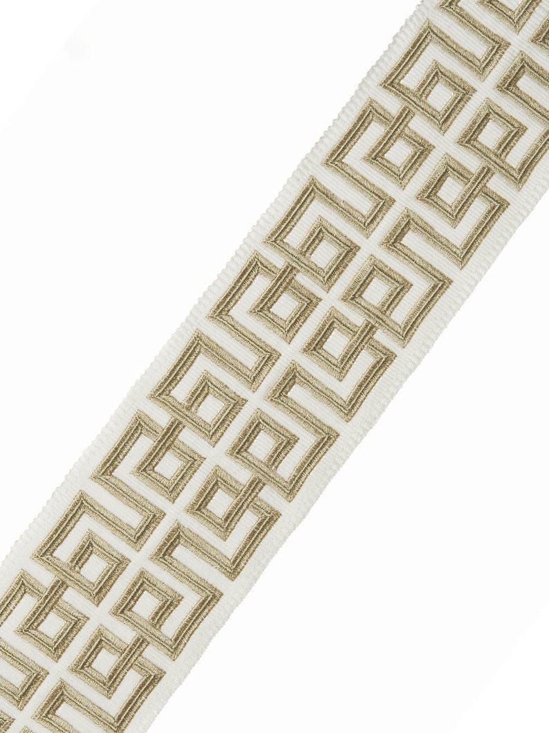 May include: White fabric trim with a gold geometric pattern. The pattern is a repeating design of squares and rectangles.