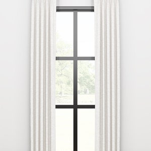 May include: A pair of white linen curtains with a subtle texture hang from a gold rod in front of a window with a black frame. The curtains are pleated and reach the floor.