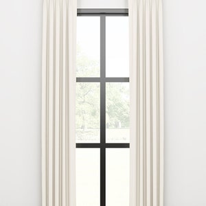 May include: Cream-colored pleated curtains hanging from a gold-colored rod, framing a black-framed window. The window offers a view of a tree and the outdoors. The curtains are floor-length and create a soft, elegant look.