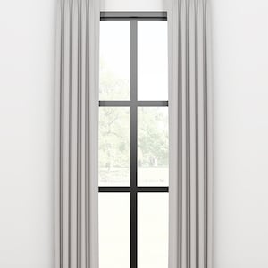 May include: A pair of white pleated curtains hanging from a gold rod in front of a window with a black frame. The curtains are drawn and the window is looking out onto a green, leafy landscape.