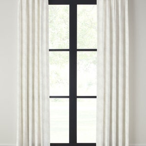May include: White linen curtains with pleats hanging from a gold rod in front of a black framed window. The curtains are drawn and the window is closed.