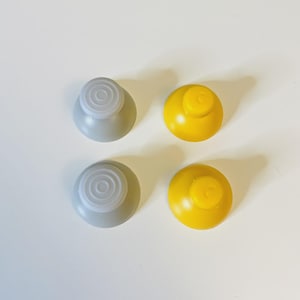 May include: Four replacement joystick thumbstick caps for a game controller. Two caps are light grey with a circular textured top surface, and two caps are solid yellow with a smooth, rounded dome shape and a small circular indentation.