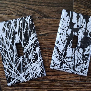 Könnte beinhalten: Zwei dekorative Lichtschalterabdeckungen mit einem abstrakten Farbspritzer-Design in Schwarz und Weiß. Die Abdeckungen haben eine rechteckige Form mit einer einzelnen Schalteröffnung und Schraubenlöchern.