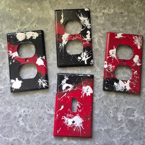May include: Four decorative electrical outlet and switch covers. The covers are painted with a black, red, and white splatter design. Two covers have two holes, and one has a single switch opening. The covers are rectangular.