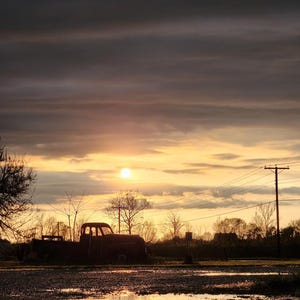 May include: A scenic sunset over a rural landscape. The silhouette of an old pickup truck is parked on a gravel lot. The sky is filled with shades of orange, yellow, and gray, with the sun setting in the distance.