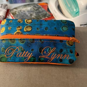 May include: A rectangular, zippered pouch with a blue background and a pattern of yellow and green circles. The pouch has an orange zipper and the name "Patty Lynn" embroidered in orange thread.
