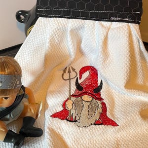 May include: White kitchen towel with a devil gnome embroidered on it. The gnome has a red hat and cape, a gray beard, and holds a trident. The towel has a black and white patterned top.
