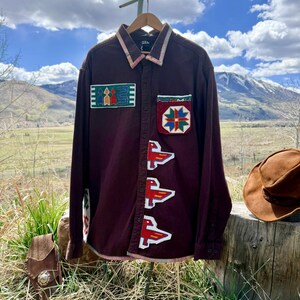 May include: A maroon button-down shirt with embroidered patches. The shirt features colourful geometric designs and red bird-like figures. The collar and pocket have patterned trim. A brown cowboy hat rests on a wooden surface in the background.