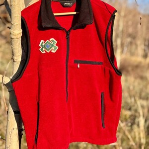 Puede incluir: Un chaleco de forro polar rojo con cuello y ribetes negros. El chaleco tiene una cremallera completa y un pequeño diseño bordado en el pecho. También tiene dos bolsillos. El chaleco está colgado de una percha de madera.