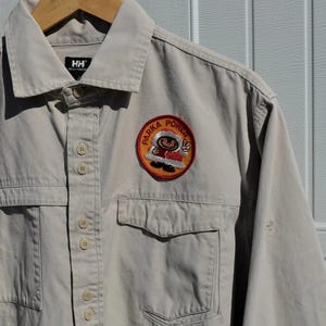 Puede incluir: Camisa beige con botones, dos bolsillos en el pecho y cuello. La camisa tiene un parche circular con la inscripción "PARKA POWER" y un personaje de dibujos animados. La camisa está colgada en una percha de madera.