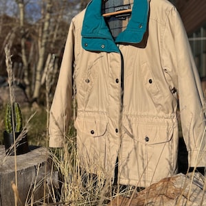 May include: Tan Pacific Trail jacket with a teal collar and plaid lining. The jacket has multiple pockets, a drawstring waist, and a button-up front. The jacket is hanging on a wooden hanger outdoors.