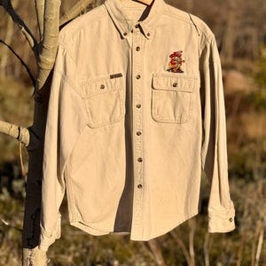 Può includere: Camicia beige con bottoni, due tasche sul petto e un piccolo disegno ricamato sulla tasca destra. La camicia ha maniche lunghe e colletto button-down. La camicia è appesa a una gruccia di legno.