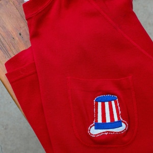 May include: A bright red ribbed knit shirt with a pocket featuring an embroidered Uncle Sam hat design. The hat has a blue brim and top, white stripes, and a white outline. The shirt has a tag that reads "Bauer 1936".