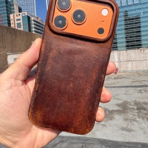 Puede incluir: Una funda de teléfono de cuero marrón con un módulo de cámara naranja. La funda tiene una textura desgastada y un ajuste perfecto. El módulo de la cámara presenta tres lentes negras y un flash blanco. La funda del teléfono se sostiene en una mano.