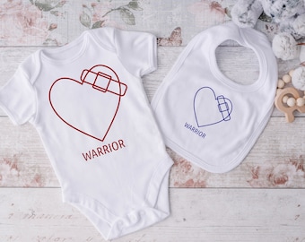 May include: White baby bodysuit and bib with a red and blue heart design. The heart has a bandage over it and the text "WARRIOR" is printed below.