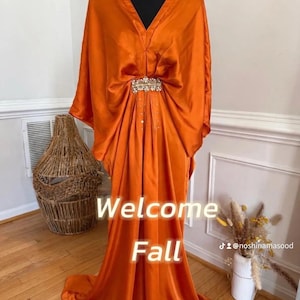 May include: A floor-length burnt orange satin kaftan dress featuring wide kimono-style sleeves, a deep V-neckline, and a central decorative silver-toned brooch with clear crystals. The text "Welcome Fall" is overlaid on the front of the garment.