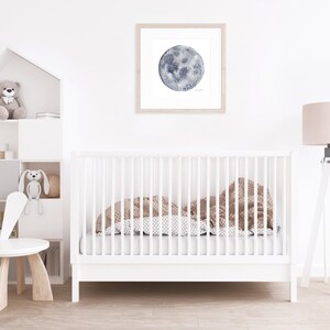 K&ouml;nnte beinhalten: Ein wei&szlig;es Babybett mit einer braunen und wei&szlig;en gemusterten Decke im Inneren. Ein wei&szlig;er Hasenstuhl steht im Vordergrund. Ein wei&szlig;es Regal mit einem Teddyb&auml;r und einem Hasenspielzeug befindet sich im Hintergrund. Ein gerahmtes Aquarellbild eines Mondes h&auml;ngt an der Wand. Eine wei&szlig;e Stehlampe mit einem beigefarbenen Lampenschirm steht in der Ecke.